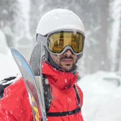 Syed Uzair Altaf in the snow, wearing a red ski jacket, white helmet and yellow goggles, with skis over his shoulder